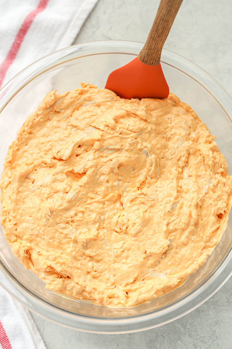 A clear glass bowl filled with creamy buffalo chicken dip with a rubber spatula leaning on the side. There is a white napkin in the background.