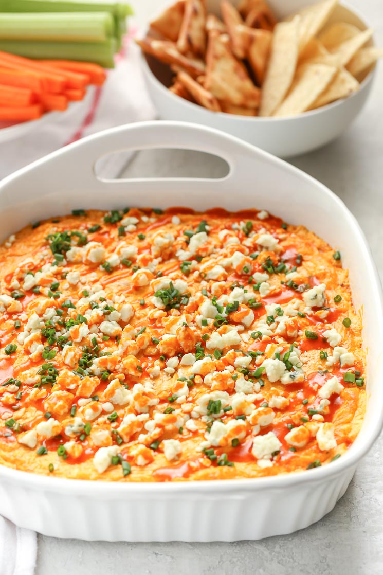 A white baking dish filled with creamy buffalo chicken dip topped with blue cheese, chives, and buffalo sauce. A bowl of chips and a plate with celery and carrot sticks in the background.