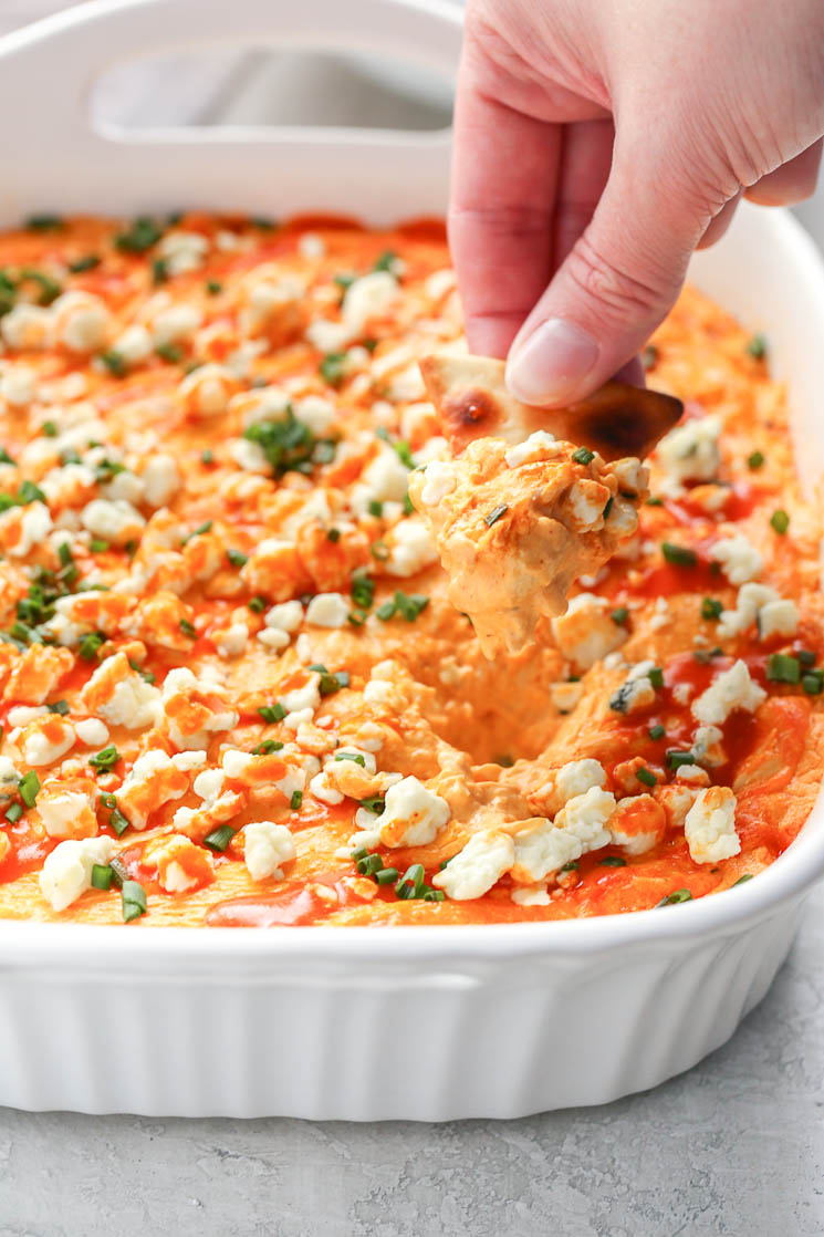 A white baking dish filled with creamy buffalo chicken dip topped with blue cheese, chives, and buffalo sauce. A hand holding a pita chip dipping into the buffalo chicken dip.