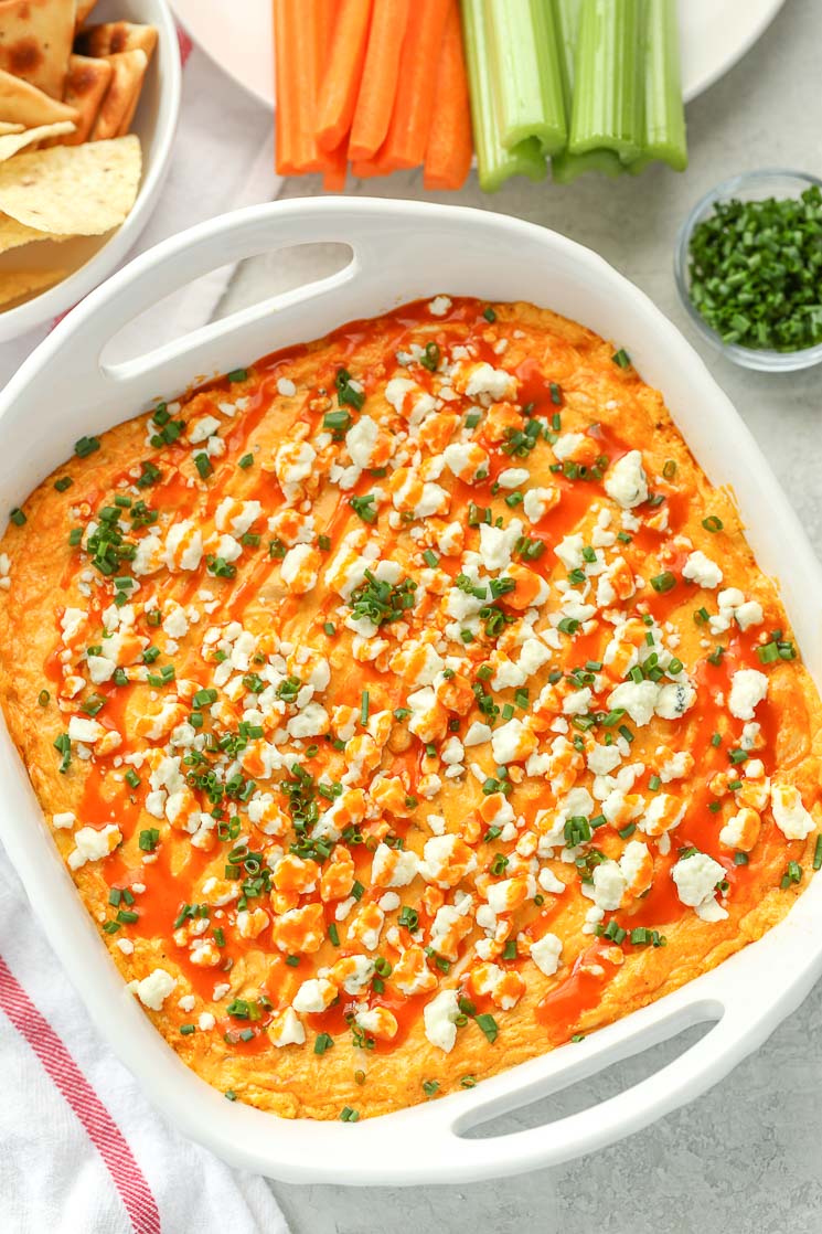 A white baking dish filled with creamy buffalo chicken dip topped with blue cheese, chives, and buffalo sauce. A bowl of chips, a plate of celery and carrot sticks and small bowl of chives around the dish.