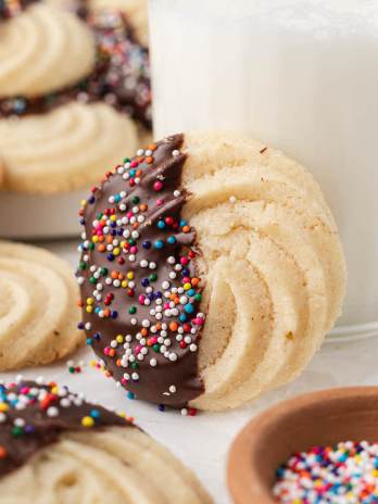 A butter cookie that has been dipped in chocolate sitting up against a glass of milk. More cookies and a bowl of sprinkles are around it.