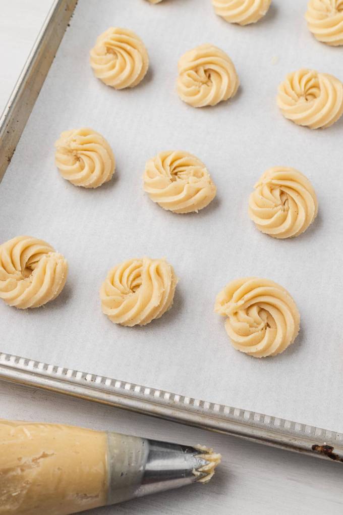 Piped butter cookie dough on a parchment-lined baking heet. A piping bag rests on the side.