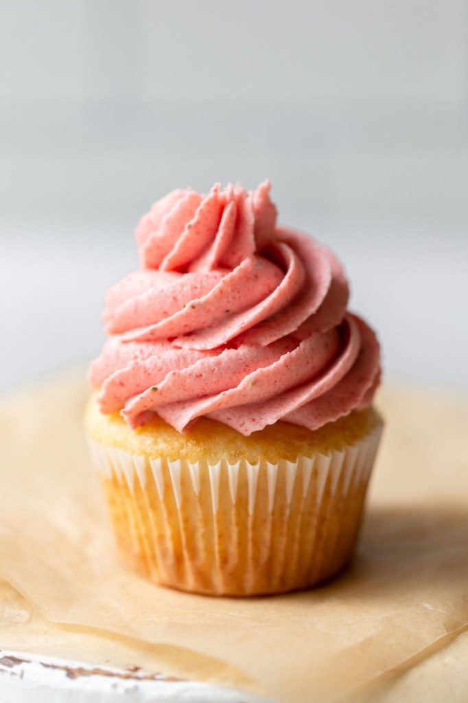 A single cupcake sitting on top of brown parchment paper topped with strawberry buttercream frosting.