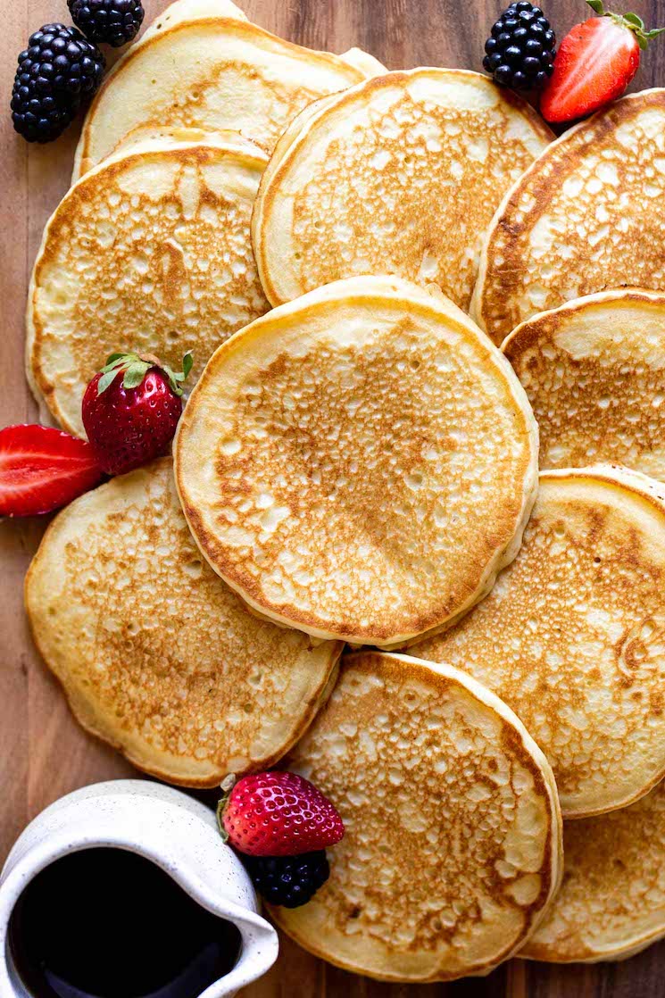 Multiple pancakes laying on a wooden surface with syrup and berries around them.