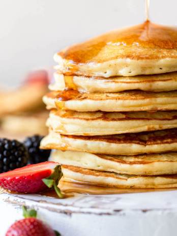 Maple syrup being poured onto a stack of pancakes on an antique white board with strawberries and blackberries surrounding it.