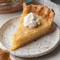 A slice of buttermilk pie topped with whipped cream on a white speckled plate.