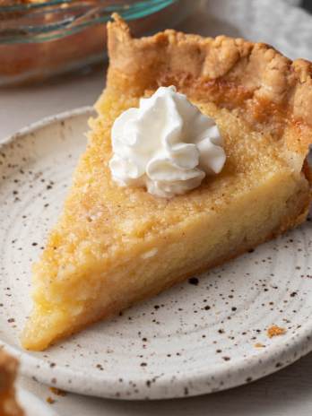 A slice of buttermilk pie topped with whipped cream on a white speckled plate.