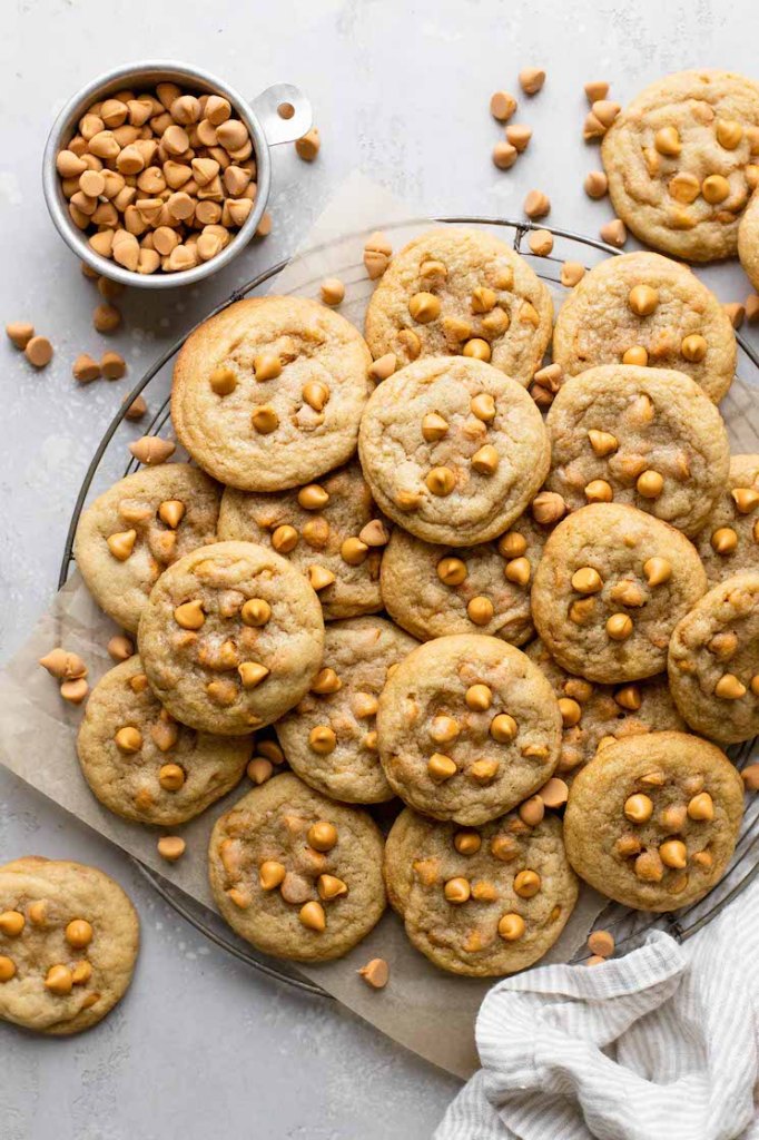 An overhead view of butterscotch cookies piled on a wire cooling rack. A dish of butterscotch chips rests on the side. 