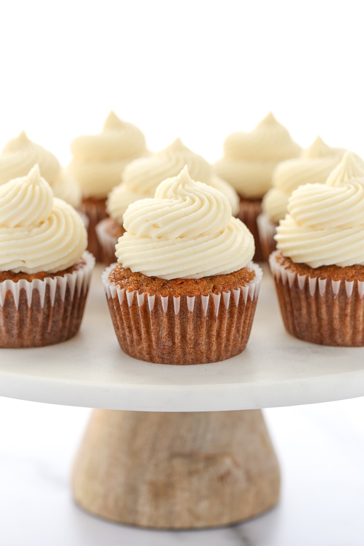A group of carrot cake cupcakes topped with cream cheese frosting resting on top of a marble cake stand with wooden base. 