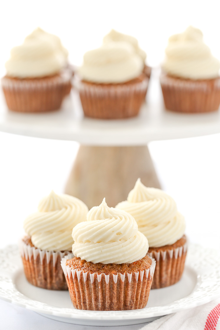 A decorative white plate holding three carrot cake cupcakes topped with cream cheese frosting in front of a marble cake stand.