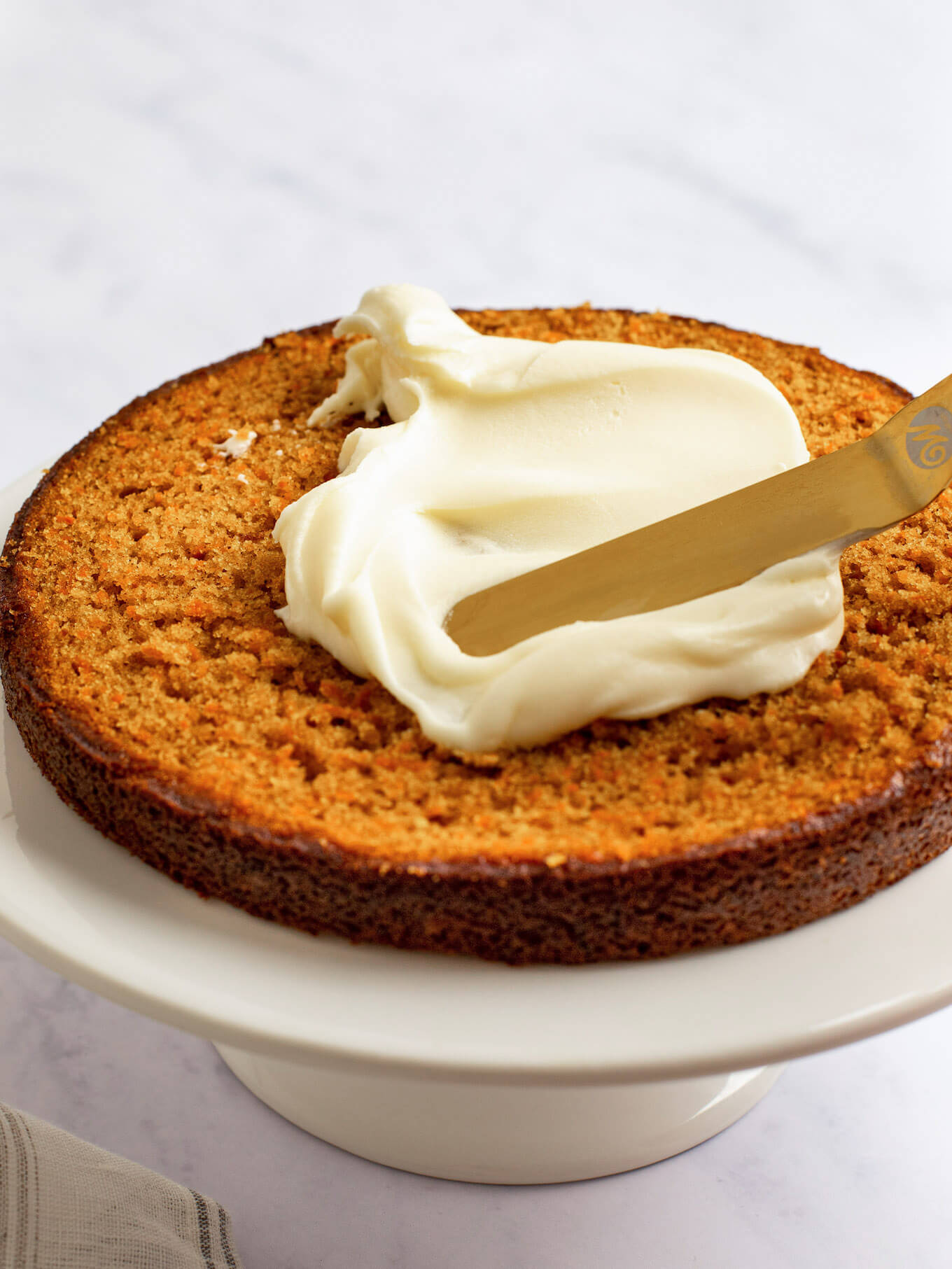 A slice of carrot cake on a cake stand. Cream cheese frosting is being spread around the cake layer.