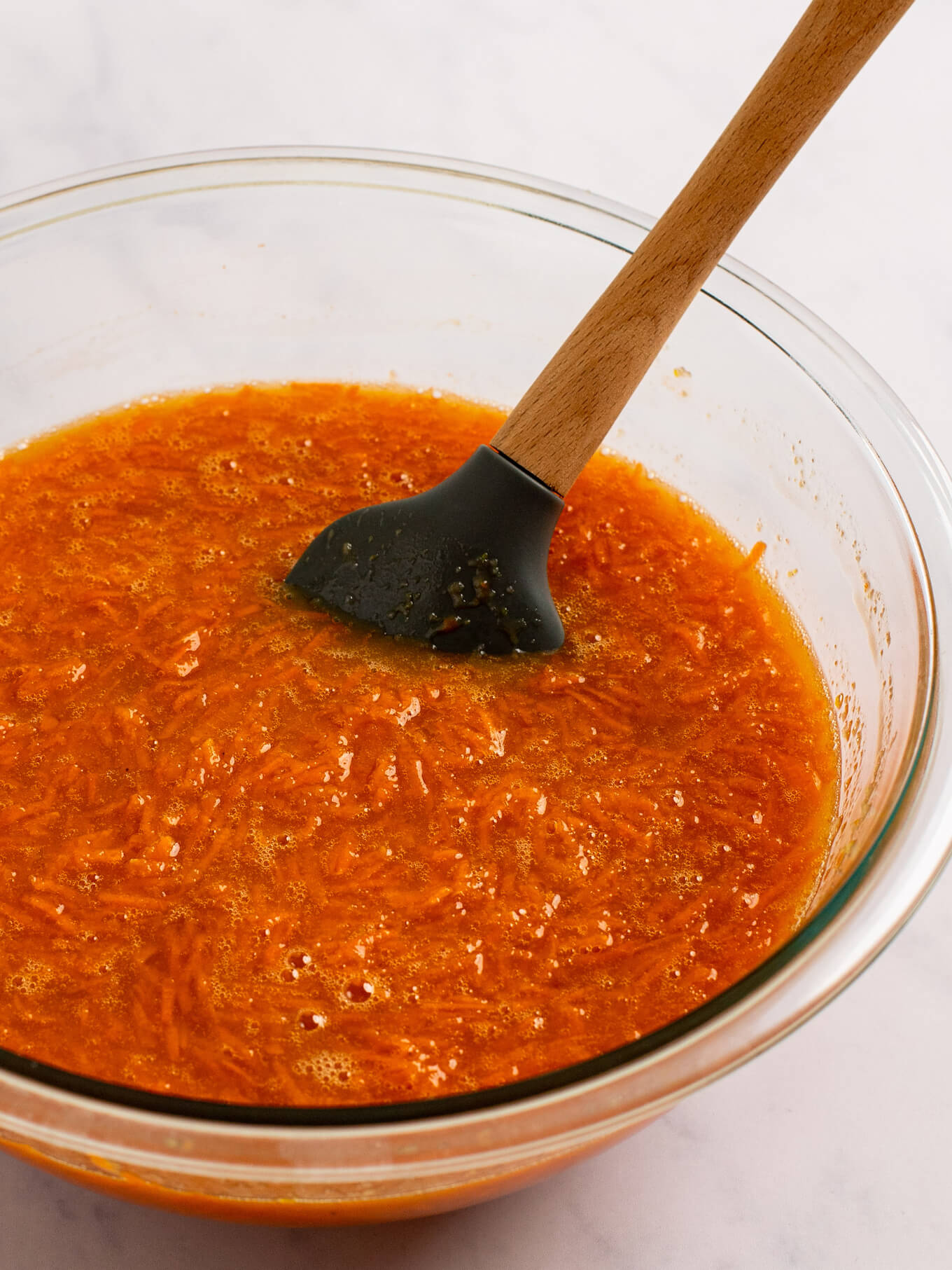 Grated carrots folded into the wet ingredients. A rubber spatula rests on the side of the bowl.