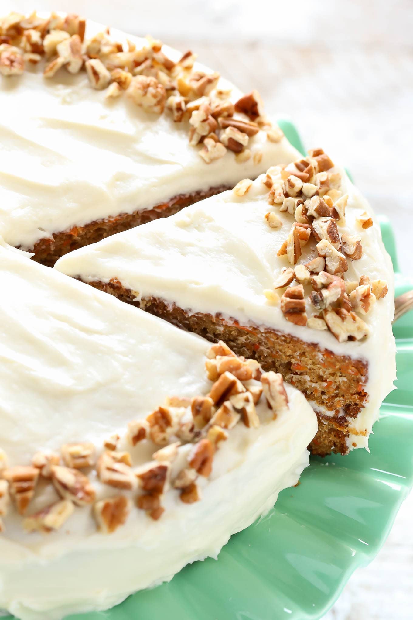 A carrot cake assembled on a green cake stand topped with cream cheese frosting and chopped pecans.