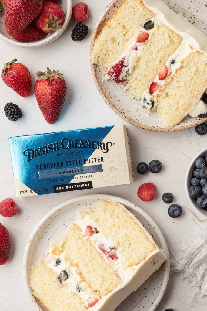 An overhead view of two slices of Chantilly cake on speckled dessert plates, with a package of Danish Creamery butter between them.