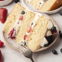 A slice of Chantilly cake on a speckle white plate. A fork rests on the plate and blueberries and strawberries are lying around it.