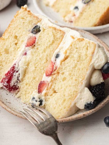 A slice of Chantilly cake on a speckle white plate. A fork rests on the plate and blueberries and strawberries are lying around it.