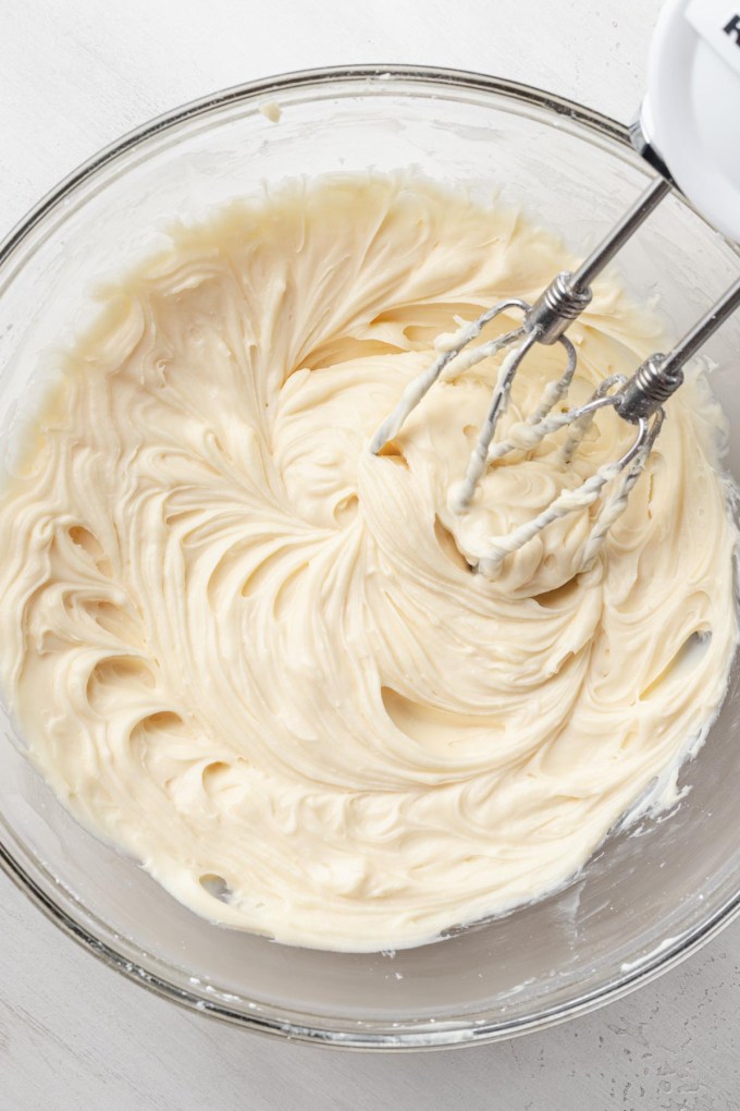 An overhead view of cream cheese being beaten in a glass mixing bowl.