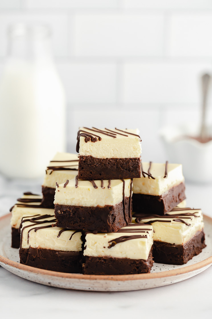 A stack of cheesecake brownies drizzled with chocolate on a rustic clay plate.