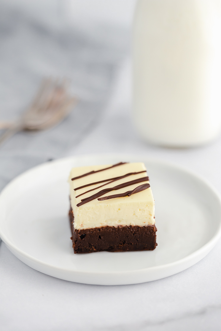 An individual cheesecake brownie drizzled with chocolate on a white plate with forks and milk in the background.