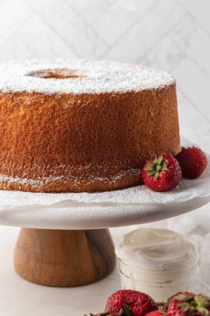 A baked chiffon cake on top of a marble cake stand. The cake has been dusted with powdered sugar and fresh strawberries are next to it.