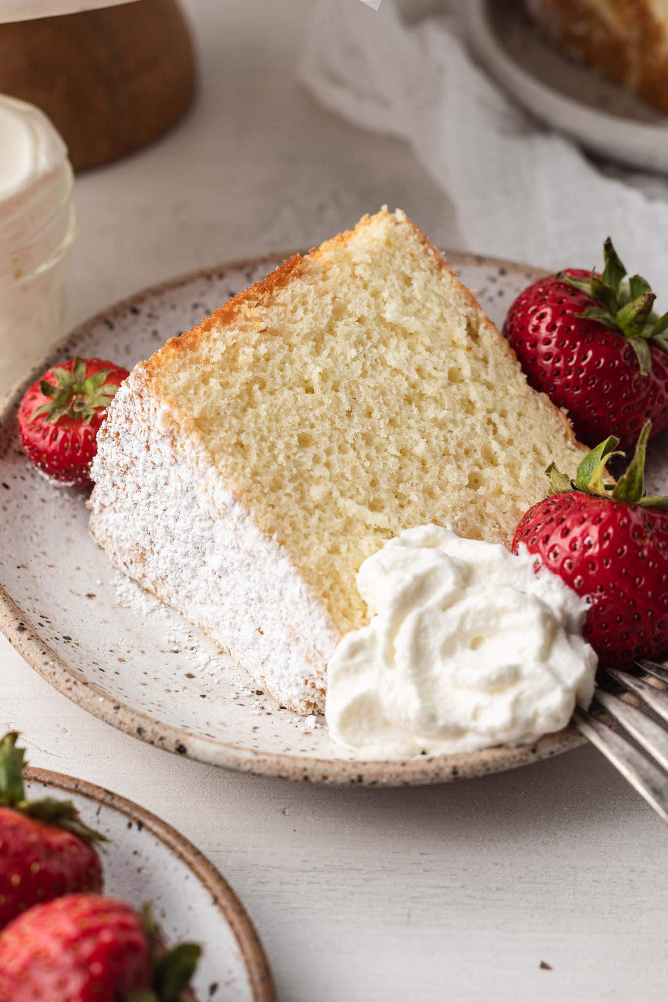 A slice of chiffon cake on a white speckled plate. The plate is garnished with fresh strawberries and a dollop of whipped cream.