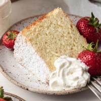 A slice of chiffon cake, fresh strawberries, and homemade whipped cream on a white speckled plate.