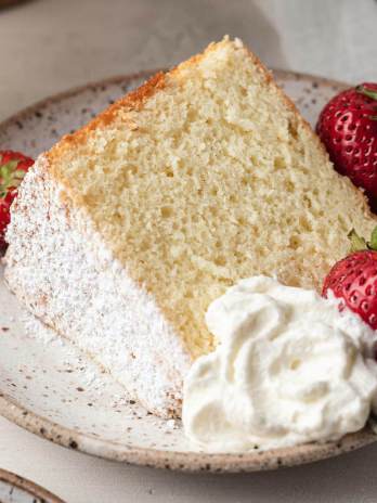 A slice of chiffon cake, fresh strawberries, and homemade whipped cream on a white speckled plate.