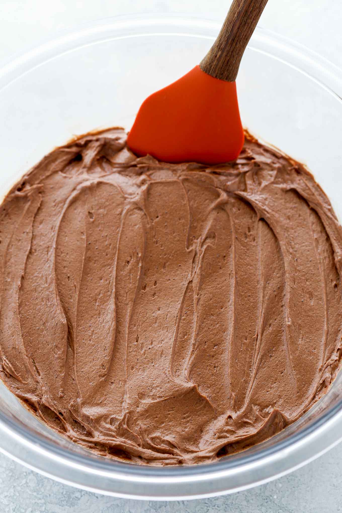 A glass bowl full of chocolate buttercream frosting with a spatula resting on the side.