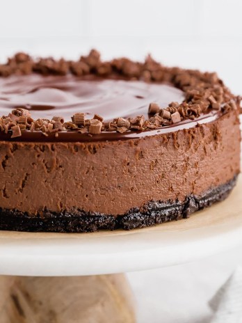 A chocolate cheesecake sitting on top of a marble cake stand. The cheesecake is topped with chocolate ganache and garnished with chocolate curls.