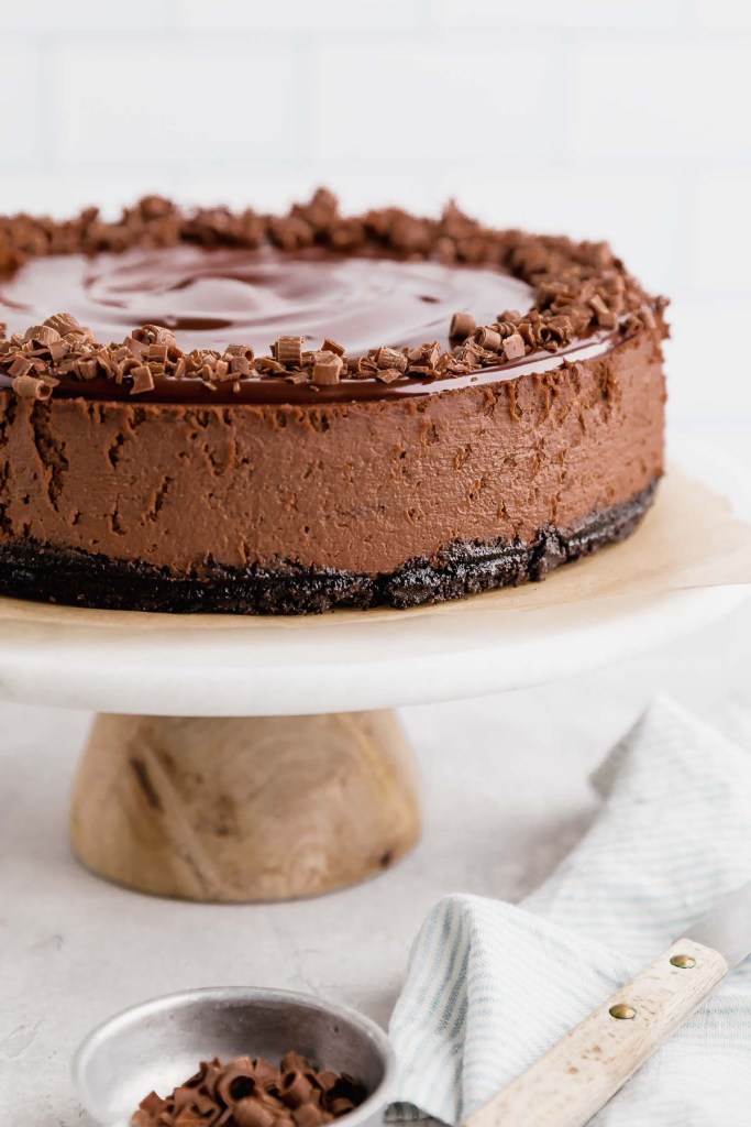 A side view of a chocolate cheesecake on a white marble cake stand. A small bowl of chocolate shavings rests in the foreground. 