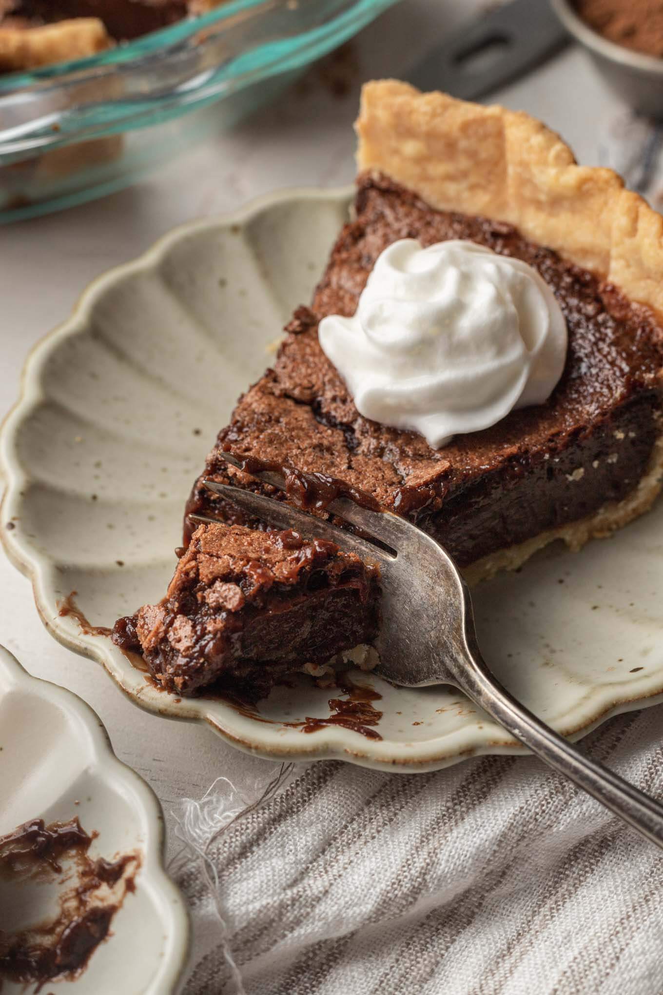 A slice of chocolate chess pie topped with whipped cream. A fork is digging into the pie. 