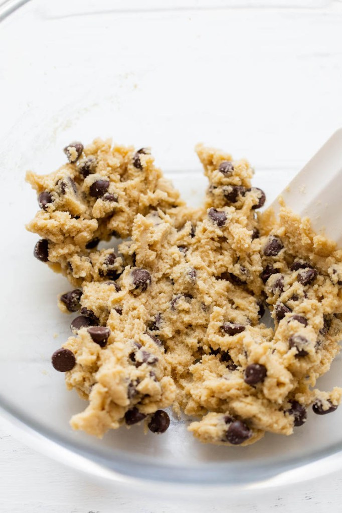 Cookie batter being mixed together with a white spatula in a clear glass bowl.