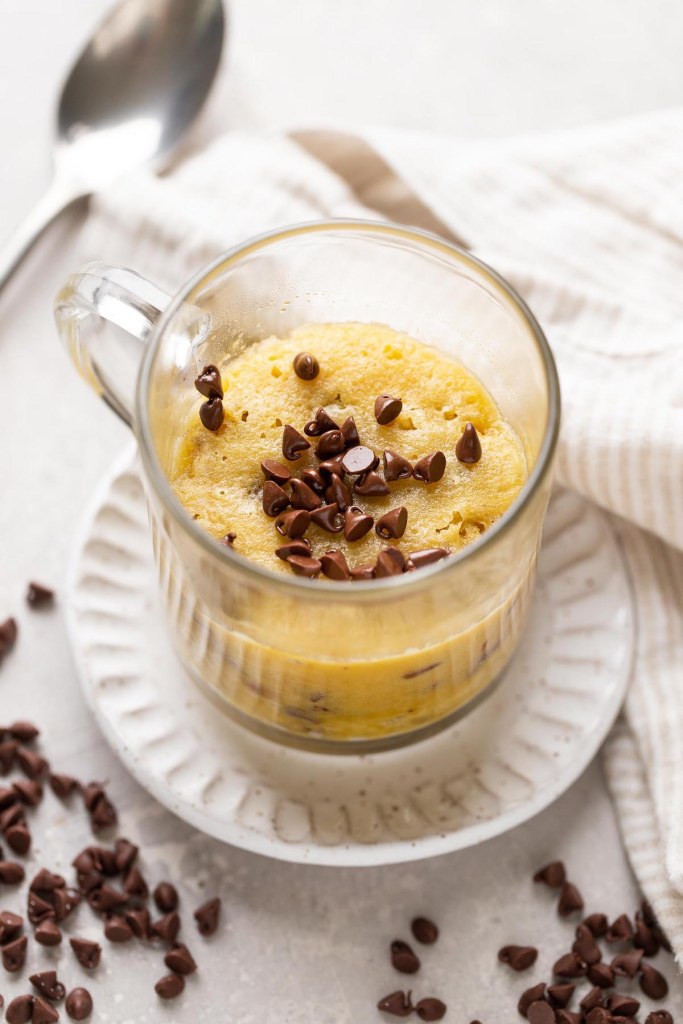 A chocolate chip cookie in a mug, on a white dessert plate. Mini chocolate chips are scattered about, and a spoon rests in the background. 