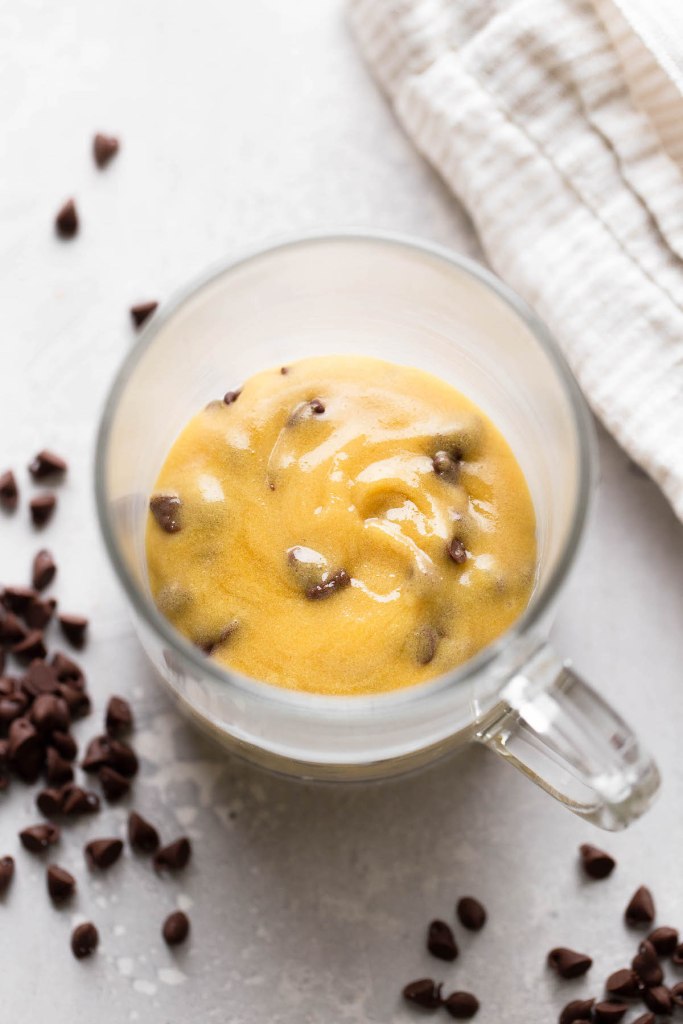 An overhead view of raw chocolate chip cookie dough in a glass mug. Mini chocolate chips are scattered around the mug. 