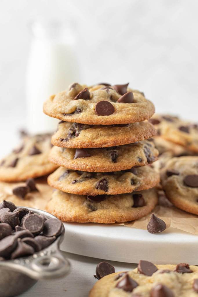 A stack of five chocolate chip pudding cookies, on a plate with more cookies. A measuring cup of chocolate chips rests in the foreground. 