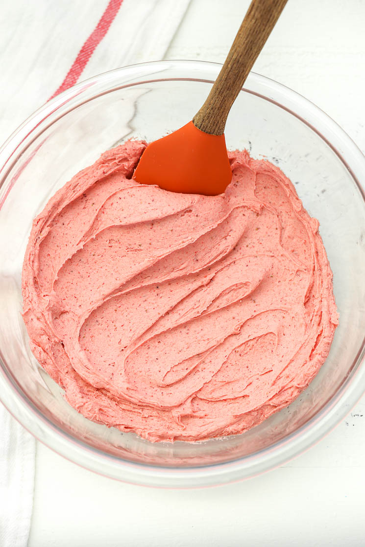 A glass bowl filled with strawberry buttercream frosting and a rubber spatula on the side.