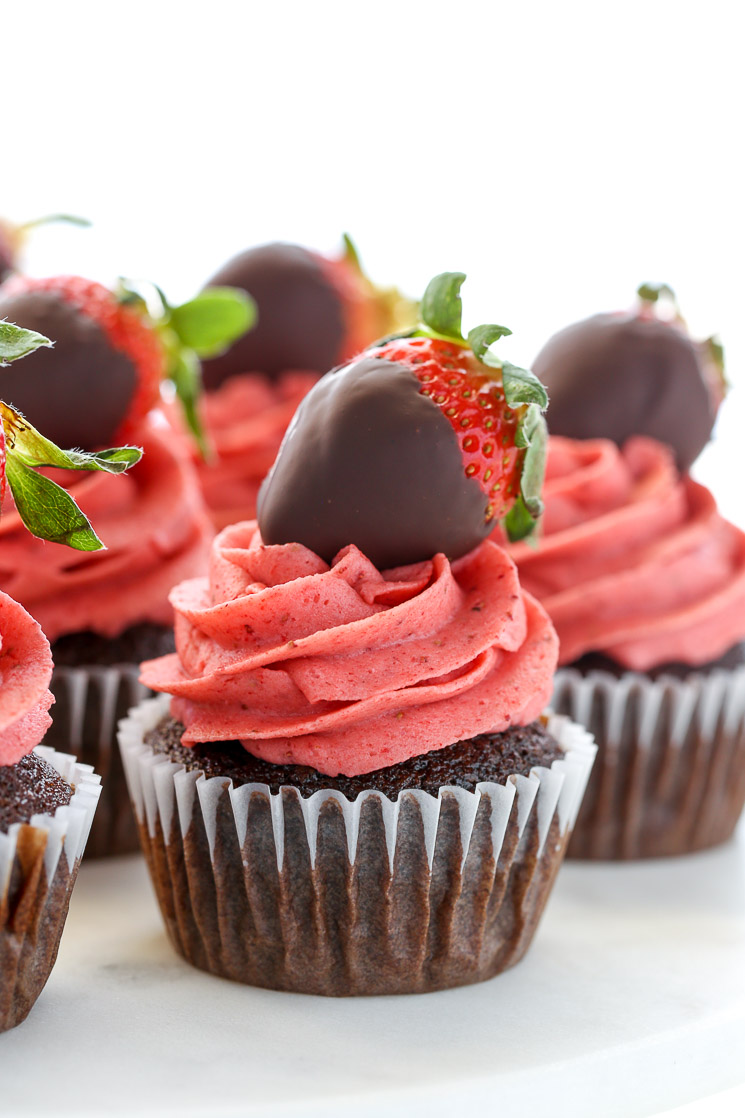 A closeup shot of chocolate cupcakes topped with strawberry buttercream frosting and chocolate covered strawberries on a white marble cake stand.