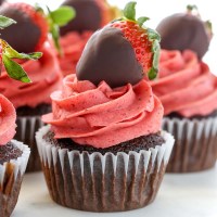 A closeup of chocolate cupcakes topped with strawberry buttercream frosting and chocolate covered strawberries.