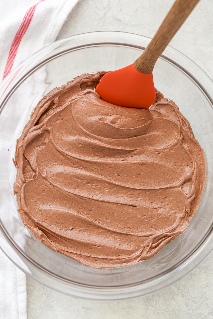 A clear glass mixing bowl filled with chocolate cream cheese frosting and a rubber spatula.