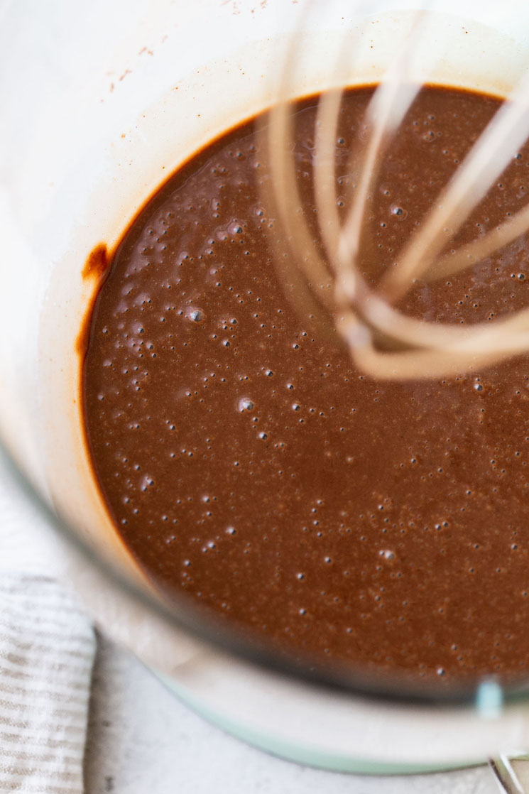 A glass mixing bowl holding chocolate cupcake batter being mixed with a whisk.