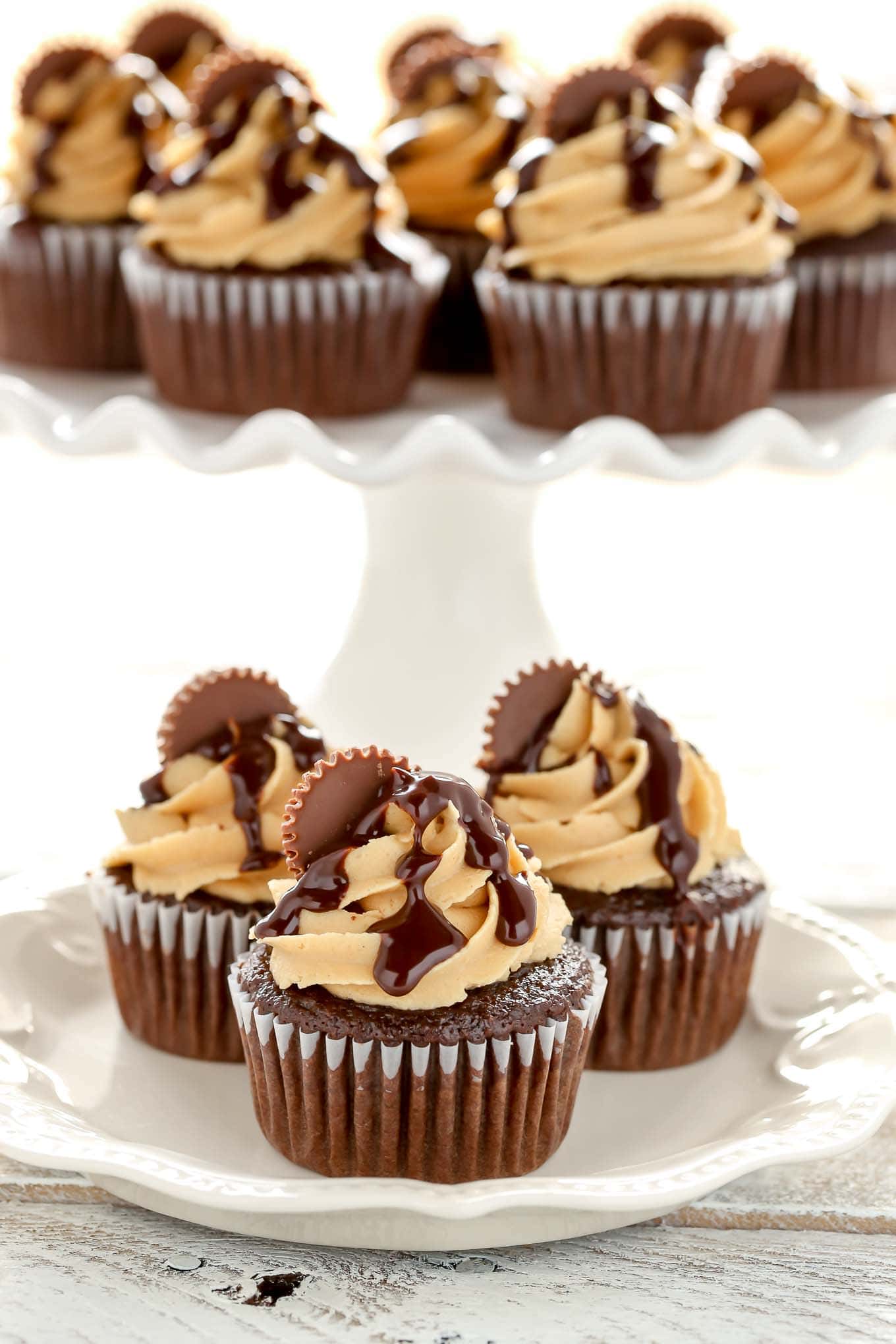 Three Reese's cupcakes on a white plate. A cake stand of cupcakes rests in the background. 