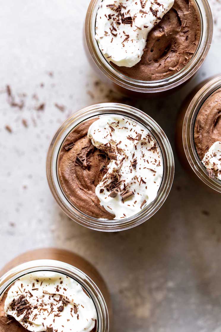 Four small glass jars filled with chocolate mousse topped with whipped cream and chocolate shaving on a gray surface dusted with more chocolate shavings.