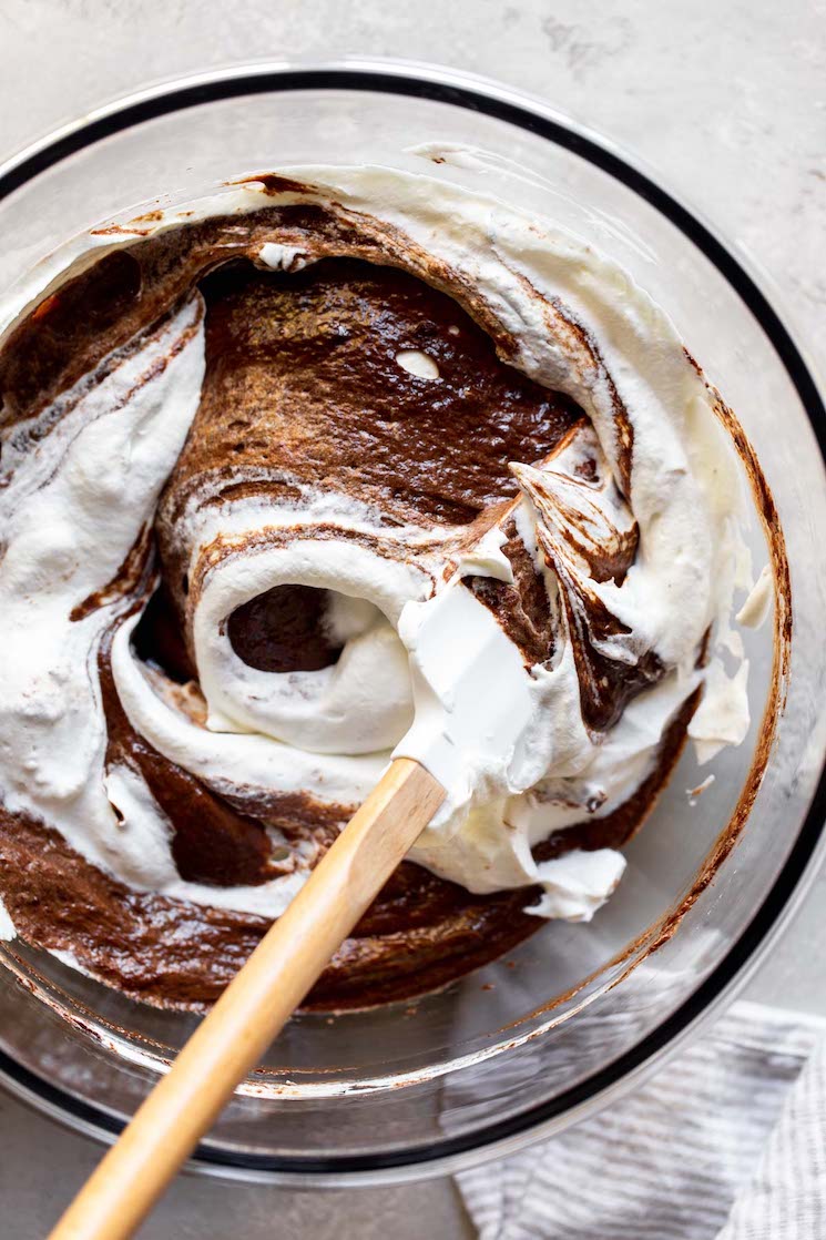 A glass mixing bowl with the chocolate mixture and whipped cream being mixed together with a rubber spatula.