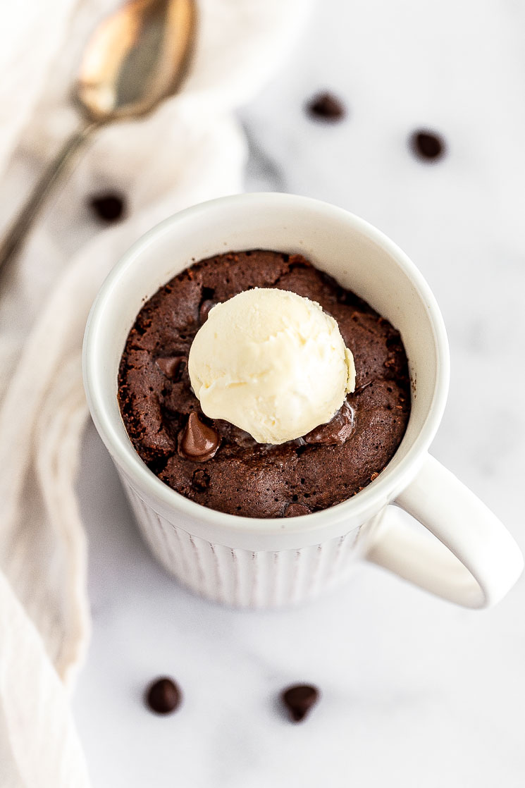 A chocolate mug cake topped with chocolate chips and a scoop of ice cream on a marble surface.
