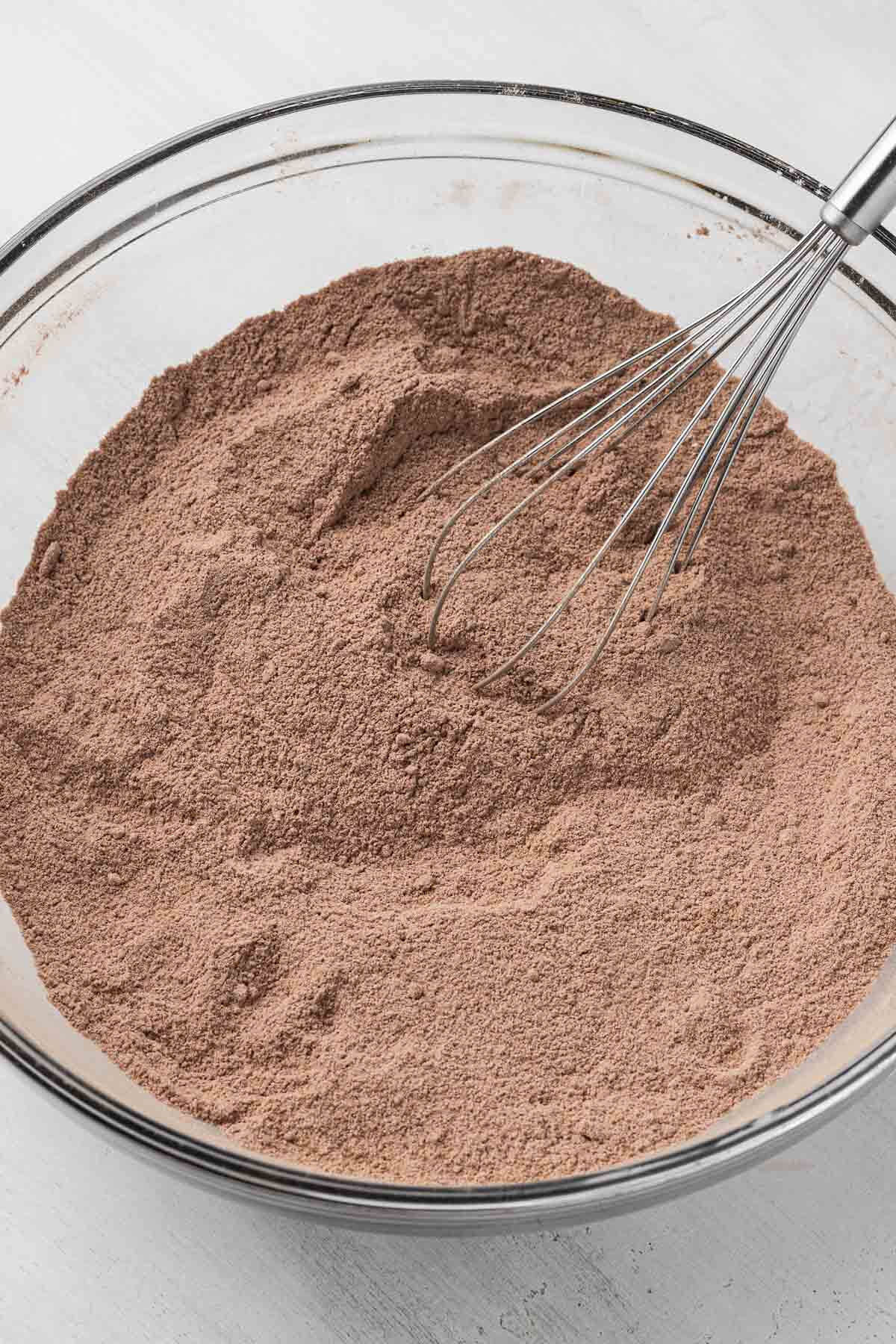 The dry ingredients for chocolate cake in a glass mixing bowl, with a whisk.