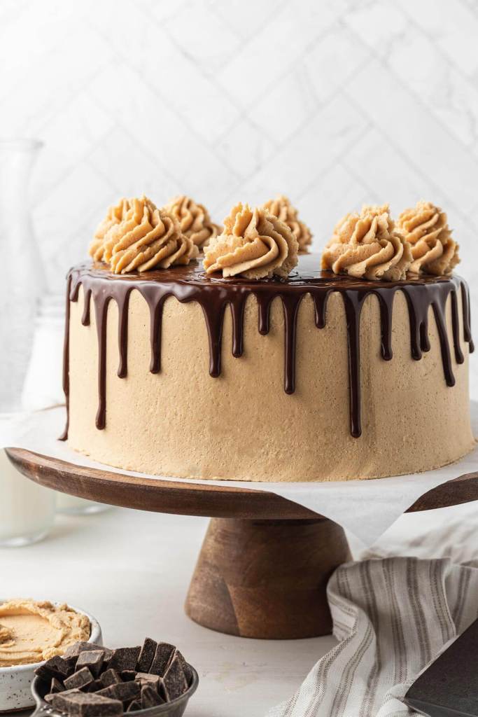 A chocolate peanut butter cake on a wooden cake stand, with chocolate chunks and peanut butter in small dishes in the foreground. 