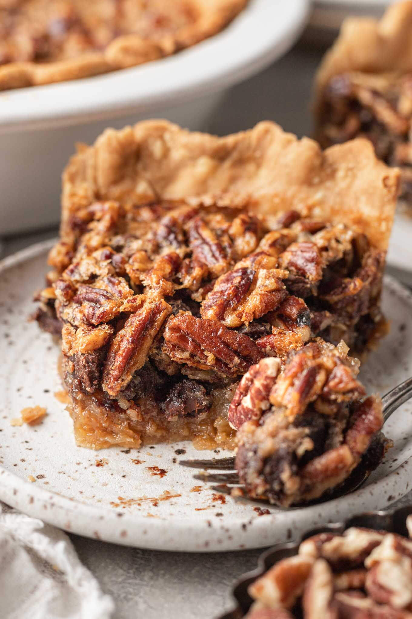 A front view of a slice of pecan chocolate pie on a speckled dessert plate. A forkful of pie rests in the foreground.  