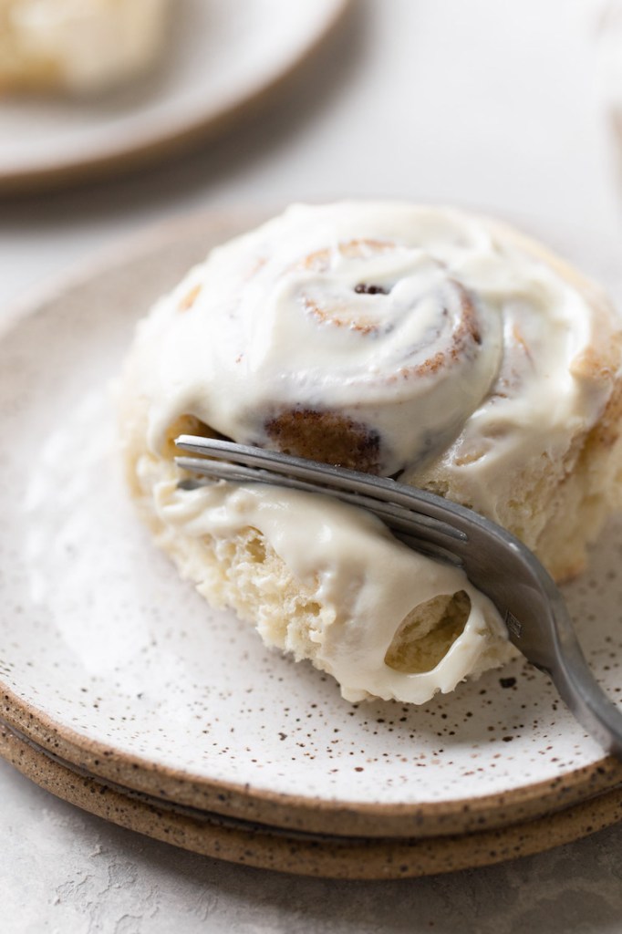 A single cinnamon roll on a white speckled plate being cut into with a fork.