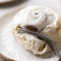 A single cinnamon roll on a white speckled plate being cut into with a fork.