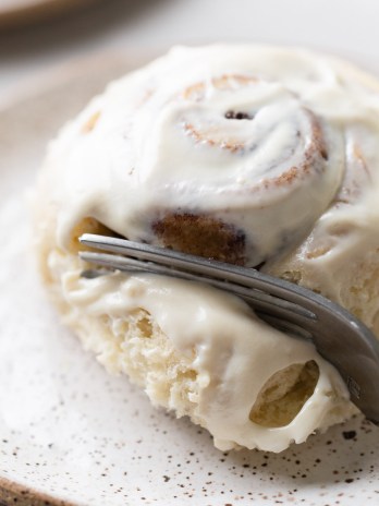 A single cinnamon roll on a white speckled plate being cut into with a fork.